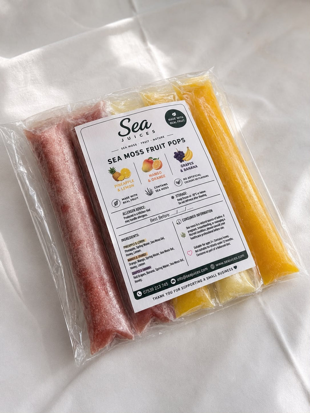 A clear plastic bag containing four frozen fruit pops in different colors, with a white product label reading 'Sea Moss Fruit Pops' and flavor icons. The pops are arranged vertically in pink, orange, yellow, and dark purple tones against a white fabric background.